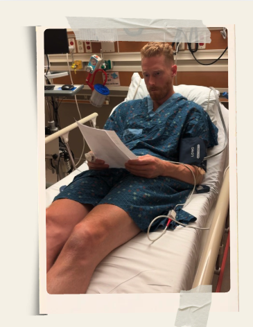 Nate in a hospital gown reading papers in an ER bed